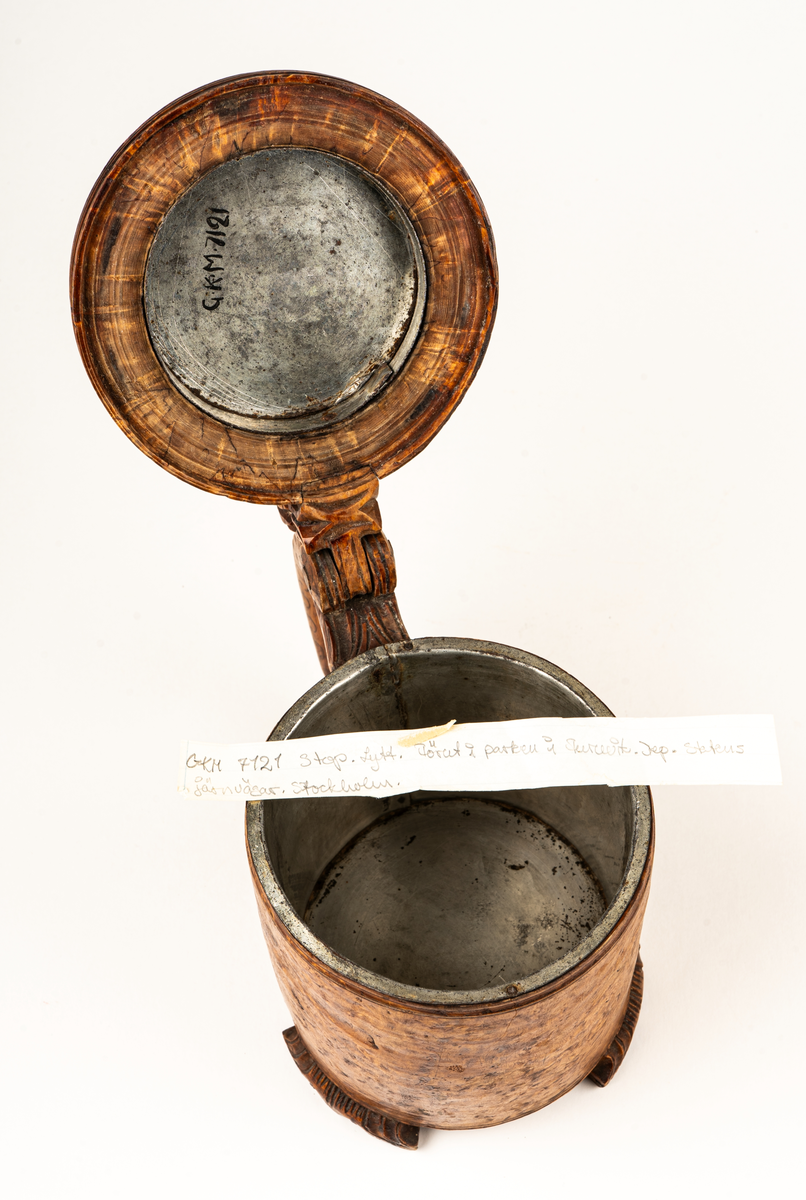 Dryckeskanna, stånka,  av masur, rundsvarvad ur ett stycke, tre låga förrer formade som blad. Knopp på handtagets övre del. På locket ristade ornament samt tjurfigur i bly. Kannan inuti plåtklädd.