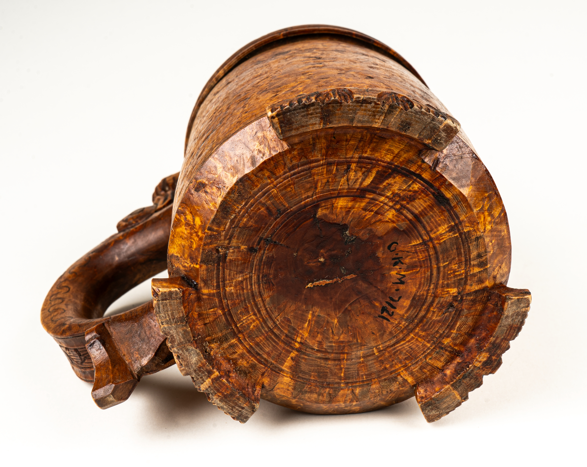 Dryckeskanna, stånka,  av masur, rundsvarvad ur ett stycke, tre låga förrer formade som blad. Knopp på handtagets övre del. På locket ristade ornament samt tjurfigur i bly. Kannan inuti plåtklädd.