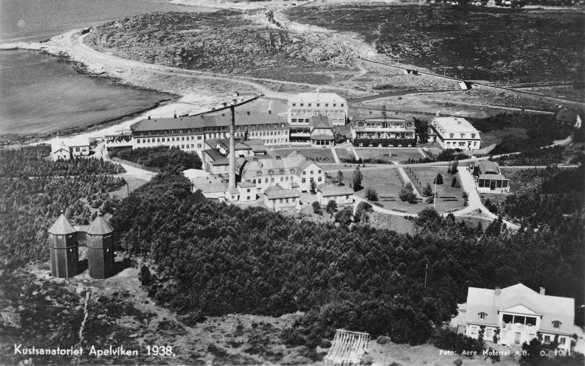 Flygbild vykort, "Kustsanatoriet Apelviken 1938." Vy över sanatorieområdet sett söderifrån med vattentornen Albert och Frida nere till vänster och överläkarvillan (Hansonska huset eller Almers hus) till vänster. Upptill ses järnvägens olika sträckningar där den först gick utmed kustlinjen runt Hästhagabergen, men från oktober 1936, i samband med järnvägens elektrifiering, drogs i rakare linje genom bergen. Den nya sträckningen går på den gamla ungefär i höjd med sanatoriet (till höger utanför bild).