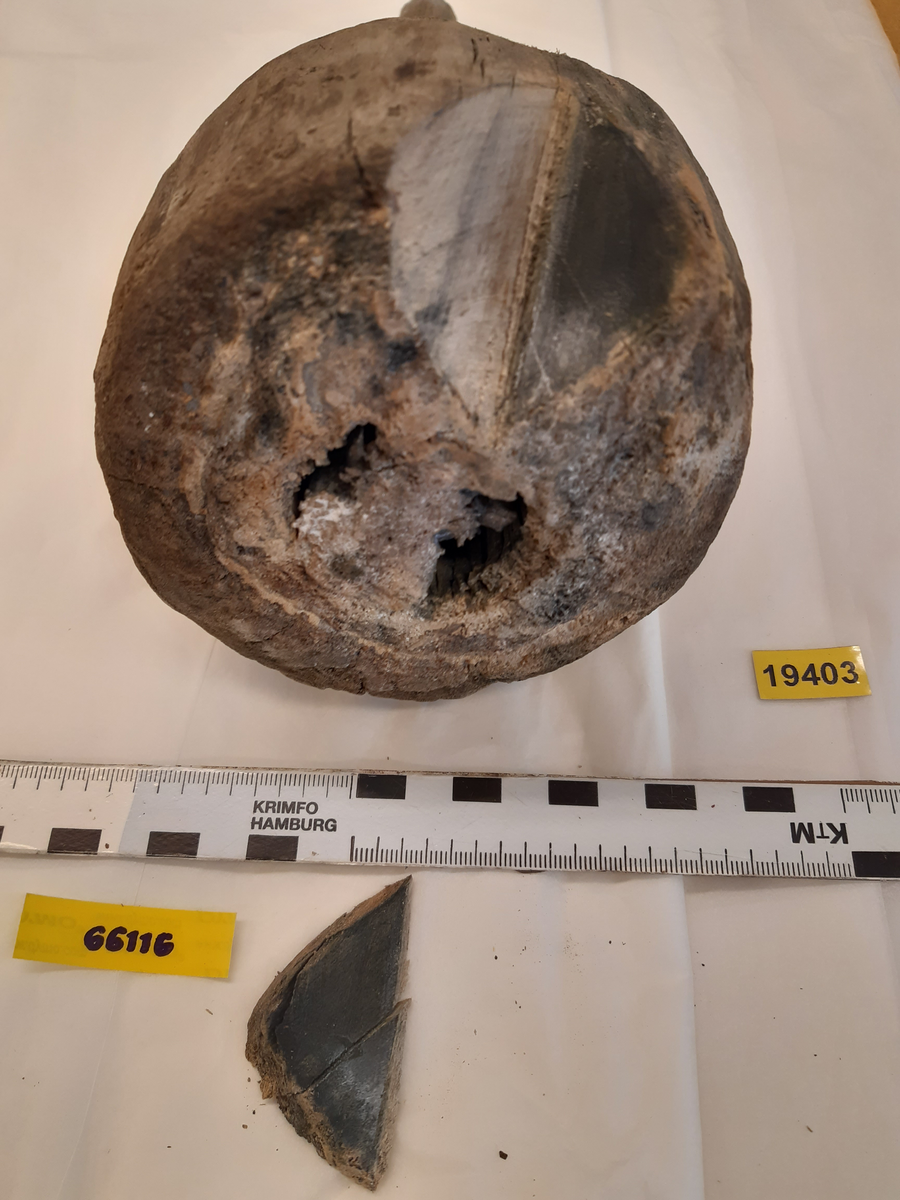 Kolv till ansättare. Kolven har ursprungligen bestått av ett svarvat trästycke med ett centrerat genomgående skafthål. Kolven har varit cylindriskt frånsett den övre änden, åt skaftet till, vilken varit profilsvarvad. Den nedre änden avslutas platt. Kolven är mycket eroderad. En del av skaftet sitter kvar i kolven.