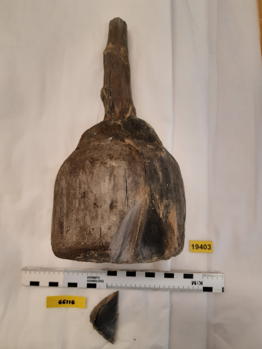 Kolv till ansättare. Kolven har ursprungligen bestått av ett svarvat trästycke med ett centrerat genomgående skafthål. Kolven har varit cylindriskt frånsett den övre änden, åt skaftet till, vilken varit profilsvarvad. Den nedre änden avslutas platt. Kolven är mycket eroderad. En del av skaftet sitter kvar i kolven.