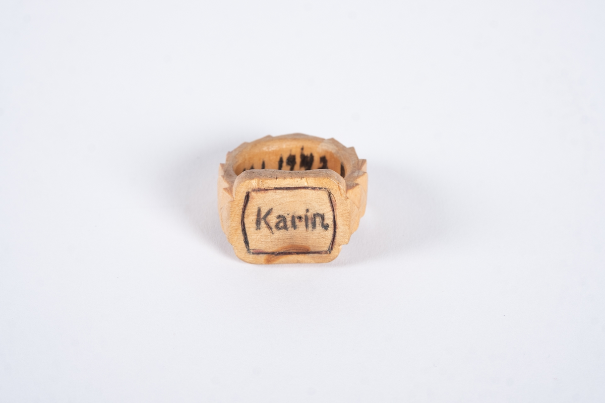 Ring av tre med utskjæringer. Det er gravert navn på ringflaten og skrift på innsiden. Ringen er lakkert.
