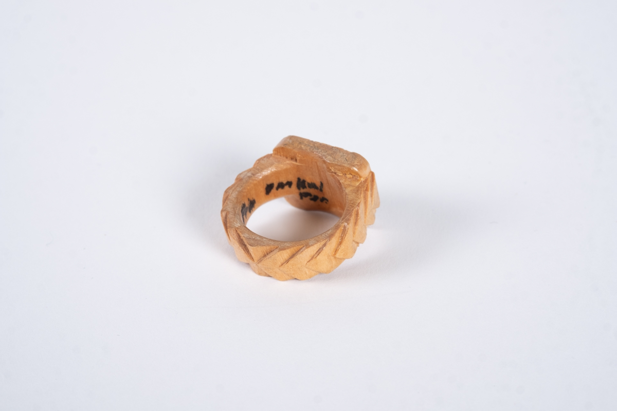Ring av tre med utskjæringer. Det er gravert navn på ringflaten og skrift på innsiden. Ringen er lakkert.