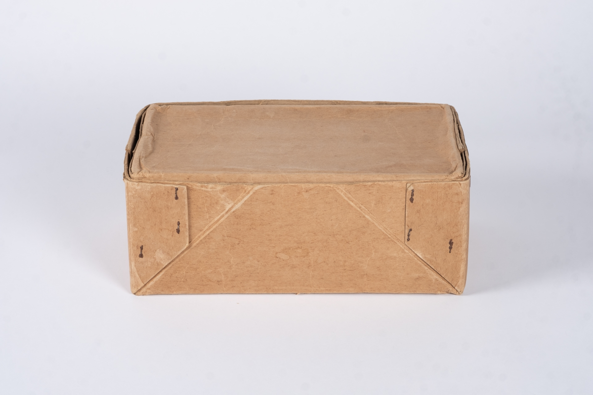 En eske av papp bestående av lokk og bunn. På oversiden av lokket er det påført tekst og logoer. I bunnen av esken ligger det tynt papir med prikker på. 