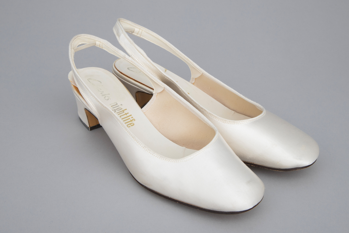 Hvite satengsko, med bred ringning, butt tå, slingback/hælstropp og kraftig, silketrukket hæl med moderat høyde. Lys brun lærlignende såle og svart gummi- eller plastlignende flikk. Ringning og slingback kantet med smalt ripsbånd og hælstroppen fôret med syntetskinn. Gulnet, kypertvevd tekstil innside. Innvendig, hvit såle av plastbelagt lerretsvev klebet på tynn papp. Den innvendige sålen er løsnet i limingen, og man kan se et løst, hælformet, hvitt papir eller vlieselinlag mellom den og selve hælen. Dette for å skjule, hindre ubehag fra de 4 spikre som hælen er festet med. Str 6, dvs 39. Ingen regulering på hælstroppen, men det er festet en flat strikk midt bak, som holder stroppen inntil hælen.