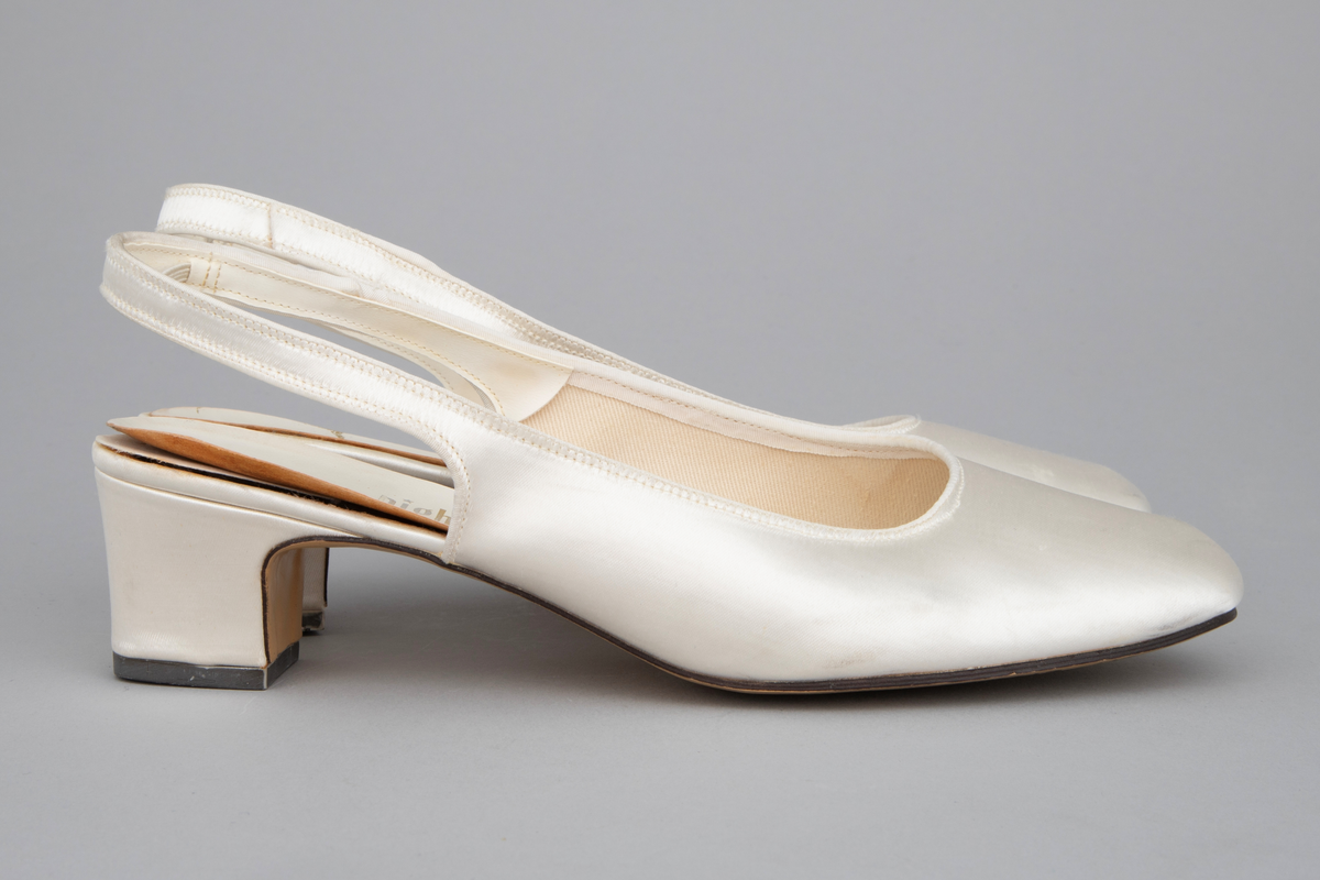 Hvite satengsko, med bred ringning, butt tå, slingback/hælstropp og kraftig, silketrukket hæl med moderat høyde. Lys brun lærlignende såle og svart gummi- eller plastlignende flikk. Ringning og slingback kantet med smalt ripsbånd og hælstroppen fôret med syntetskinn. Gulnet, kypertvevd tekstil innside. Innvendig, hvit såle av plastbelagt lerretsvev klebet på tynn papp. Den innvendige sålen er løsnet i limingen, og man kan se et løst, hælformet, hvitt papir eller vlieselinlag mellom den og selve hælen. Dette for å skjule, hindre ubehag fra de 4 spikre som hælen er festet med. Str 6, dvs 39. Ingen regulering på hælstroppen, men det er festet en flat strikk midt bak, som holder stroppen inntil hælen.