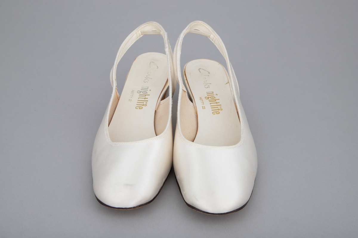 Hvite satengsko, med bred ringning, butt tå, slingback/hælstropp og kraftig, silketrukket hæl med moderat høyde. Lys brun lærlignende såle og svart gummi- eller plastlignende flikk. Ringning og slingback kantet med smalt ripsbånd og hælstroppen fôret med syntetskinn. Gulnet, kypertvevd tekstil innside. Innvendig, hvit såle av plastbelagt lerretsvev klebet på tynn papp. Den innvendige sålen er løsnet i limingen, og man kan se et løst, hælformet, hvitt papir eller vlieselinlag mellom den og selve hælen. Dette for å skjule, hindre ubehag fra de 4 spikre som hælen er festet med. Str 6, dvs 39. Ingen regulering på hælstroppen, men det er festet en flat strikk midt bak, som holder stroppen inntil hælen.