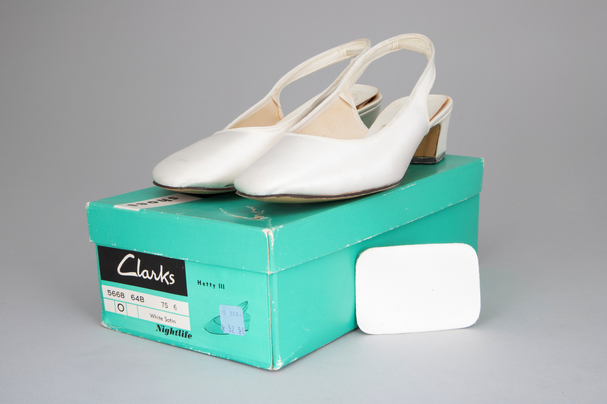 Hvite satengsko, med bred ringning, butt tå, slingback/hælstropp og kraftig, silketrukket hæl med moderat høyde. Lys brun lærlignende såle og svart gummi- eller plastlignende flikk. Ringning og slingback kantet med smalt ripsbånd og hælstroppen fôret med syntetskinn. Gulnet, kypertvevd tekstil innside. Innvendig, hvit såle av plastbelagt lerretsvev klebet på tynn papp. Den innvendige sålen er løsnet i limingen, og man kan se et løst, hælformet, hvitt papir eller vlieselinlag mellom den og selve hælen. Dette for å skjule, hindre ubehag fra de 4 spikre som hælen er festet med. Str 6, dvs 39. Ingen regulering på hælstroppen, men det er festet en flat strikk midt bak, som holder stroppen inntil hælen.
