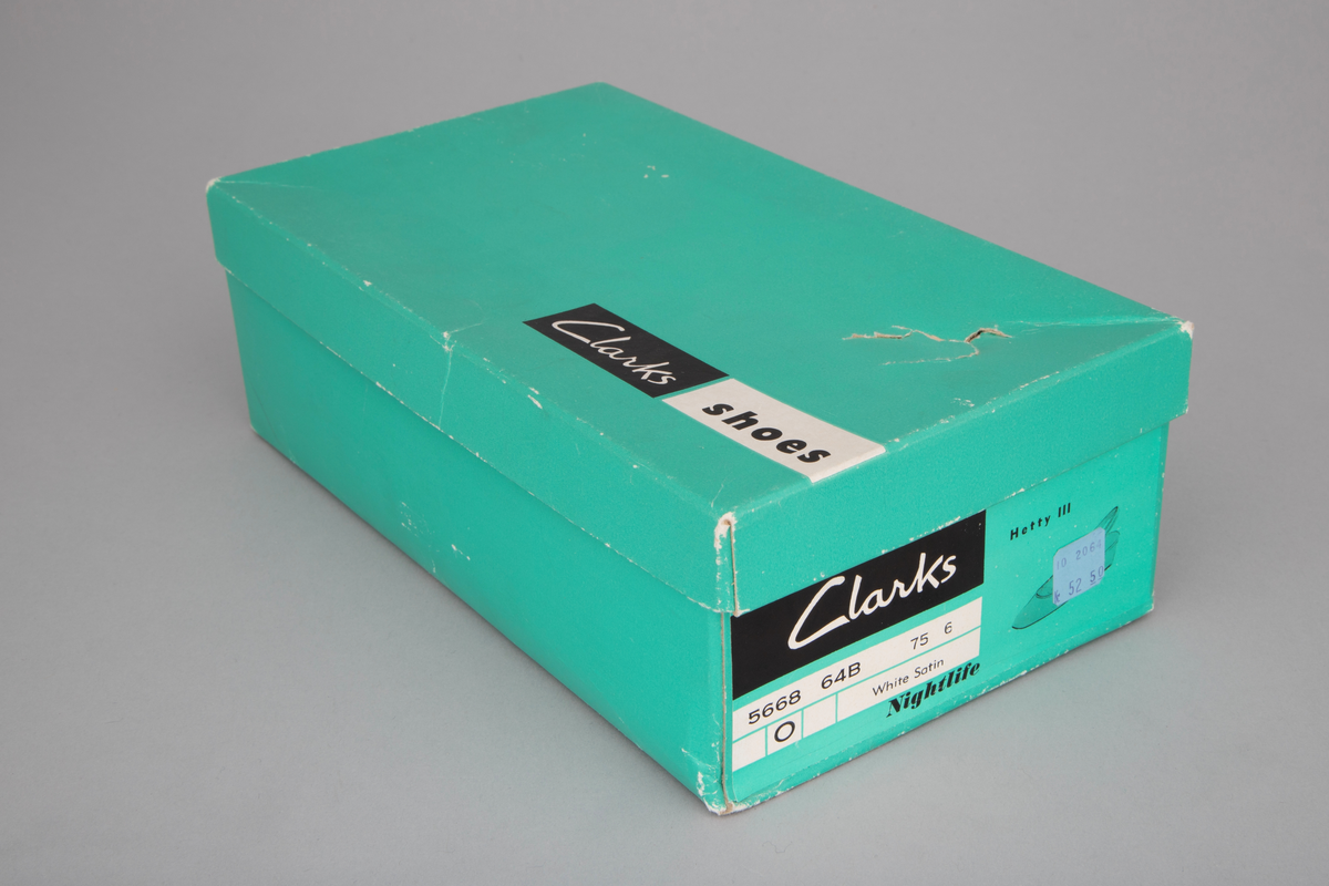 Hvite satengsko, med bred ringning, butt tå, slingback/hælstropp og kraftig, silketrukket hæl med moderat høyde. Lys brun lærlignende såle og svart gummi- eller plastlignende flikk. Ringning og slingback kantet med smalt ripsbånd og hælstroppen fôret med syntetskinn. Gulnet, kypertvevd tekstil innside. Innvendig, hvit såle av plastbelagt lerretsvev klebet på tynn papp. Den innvendige sålen er løsnet i limingen, og man kan se et løst, hælformet, hvitt papir eller vlieselinlag mellom den og selve hælen. Dette for å skjule, hindre ubehag fra de 4 spikre som hælen er festet med. Str 6, dvs 39. Ingen regulering på hælstroppen, men det er festet en flat strikk midt bak, som holder stroppen inntil hælen.