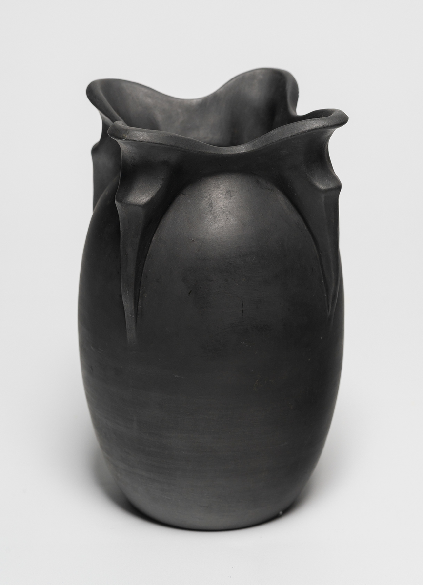 Vase i sortbrent leirgods. Eggeformet korpus med organisk munningsrand, i form av fire utoverbøyde plastiske tunger. Matt ytterside og plan bunn.