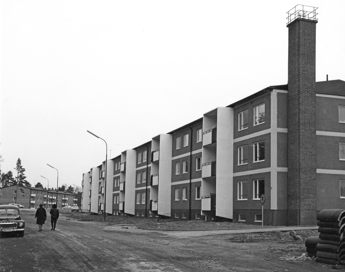 Nybyggda flerfamiljhus på Estövägen, kvarteret Duvhöken. Två personer går längs den ännu inte asfalterade vägen. 

Bebyggelse, trafik.
