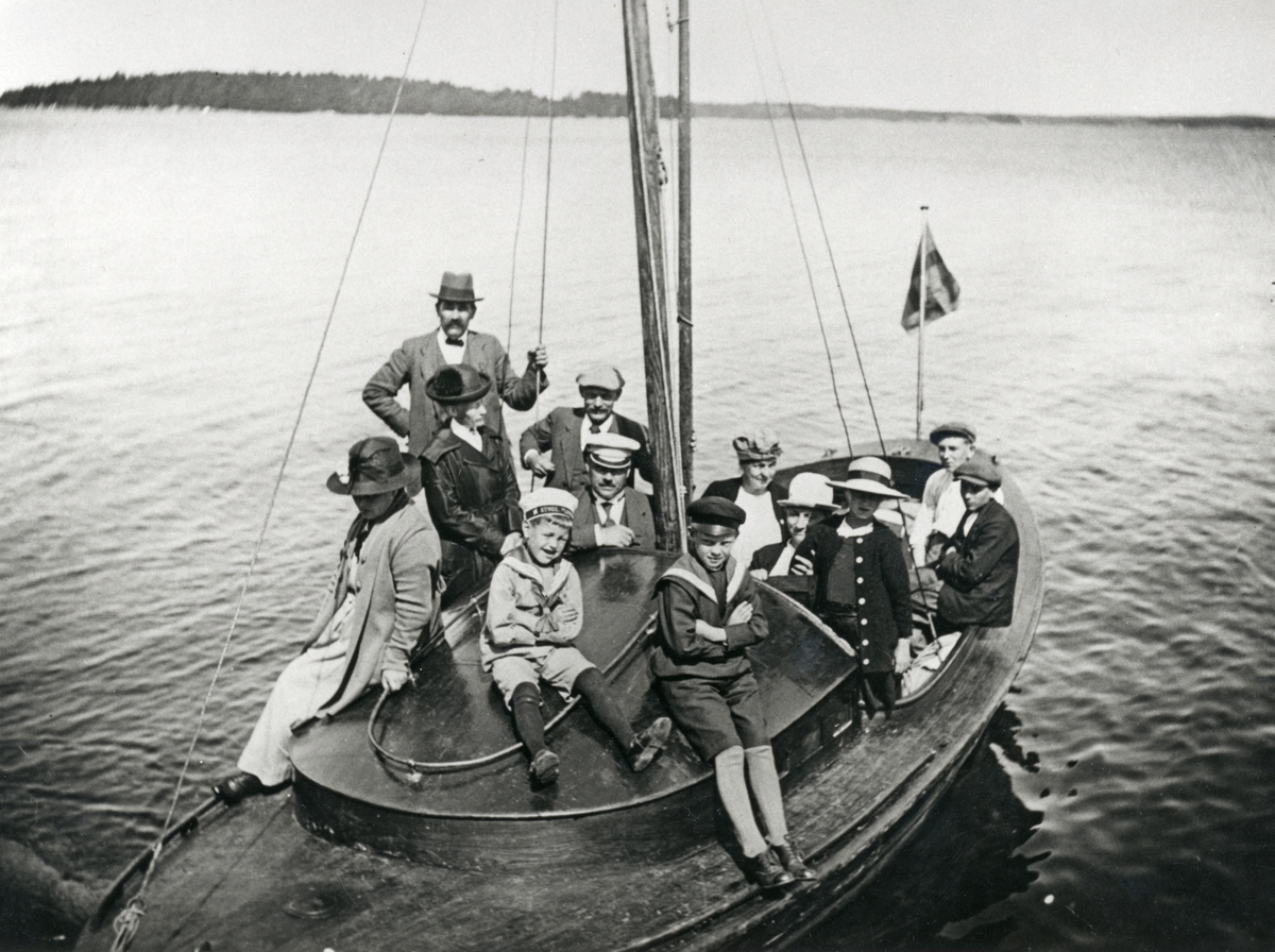 Båtutflykt, en grupp finklädda män, kvinnor och barn sitter i en träbåt. 

Sjöfart, fritid, sommar.