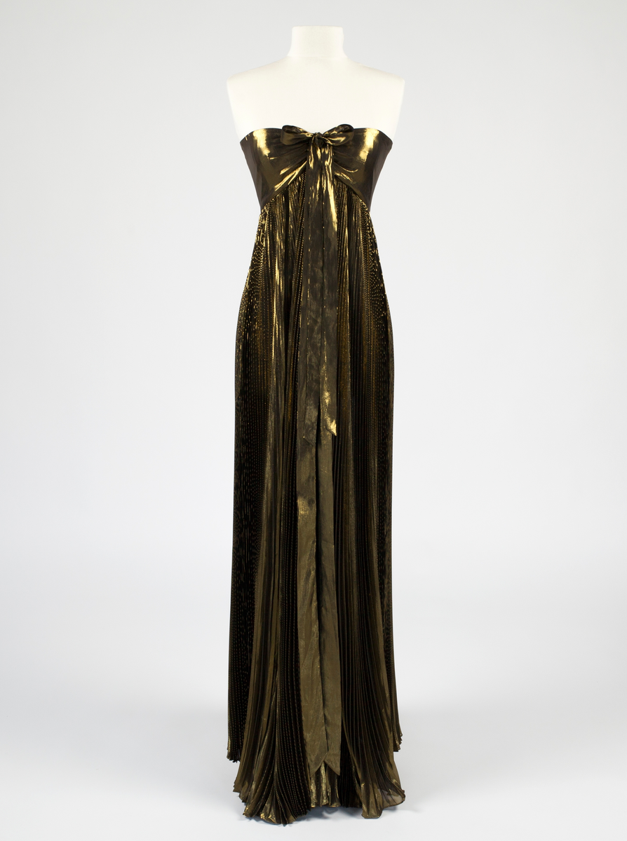 Fotsid kjole med slep. Overdelen er tettsittende og stroppeløs, holdt oppe med 10 spiler mellom det metallinnvirkede, bronsefargede stoffet og det sorte fôrstoffet. Frontstykket er rynket sammen mellom brystene og prydet med to korte og to lange bånd av kjolestoffet, de to korte antagelig ment å bli knyttet rundt nakken eller som sløyfe, mens de to lange skal henge fritt. Bakstykket er lavt og rett skåret i overkant, i høyde med bryst og ermehule. Nederst er livstykket skåret ned i spiss midt bak. Kjolen lukkes midt bak med glidelås og hekte øverst. Fra midjesømmen flommer et plissert skjørt ut i stor vidde med slep.