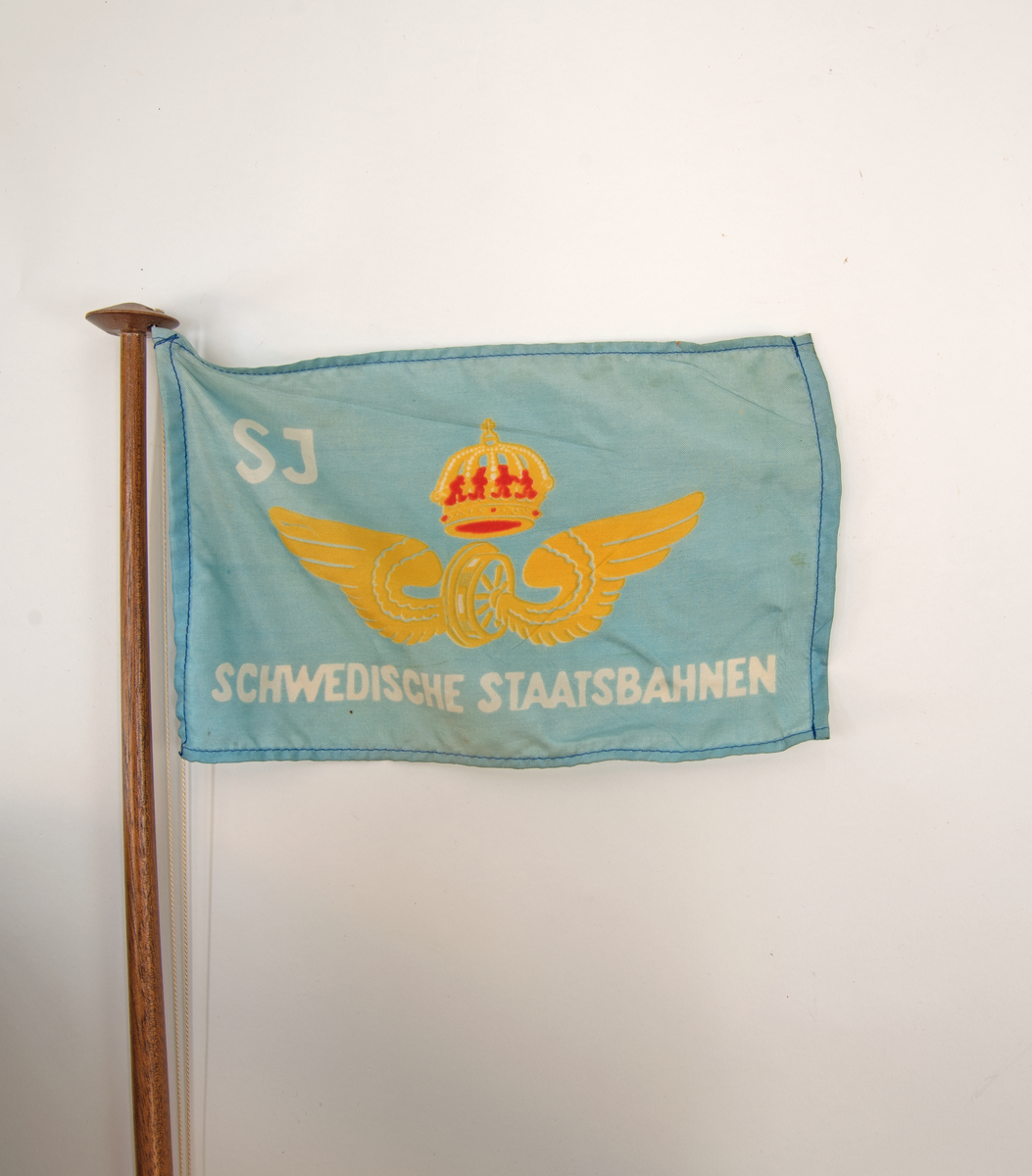 Bordstandar med blå flagga med SJ:s logotyp och texten: "SCHWEDISCHE STAATSBAHNEN". Stång och fot av trä, på foten tryckt text: "SJ OSTSEE - LINIEN" "TRAVEMÜNDE - MALMÖ".