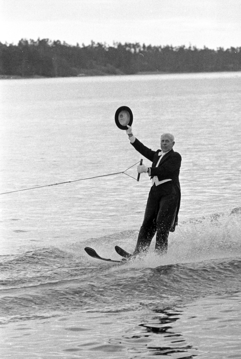 Nynäshamns Vattenskidklubbs årliga show i Nynäshamns hamn på nyårsdagen. Den snart 80-åriga Ragnar Frunck åker vattenskidor i frack och lyfter på hatten för publiken. Fotografiet publicerat i Nynäshamns Posten 1974-01-04. 

Sport, fritid, nöje, underhållning.

Personer på bilden:
Ragnar Frunck