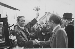 En mann som håndhilser på en annen med hatt. Den første mannen holder en blomsterbukett opp i været med sin venstre hånd. En stor, smilende folkemengde sees i bakgrunnen.