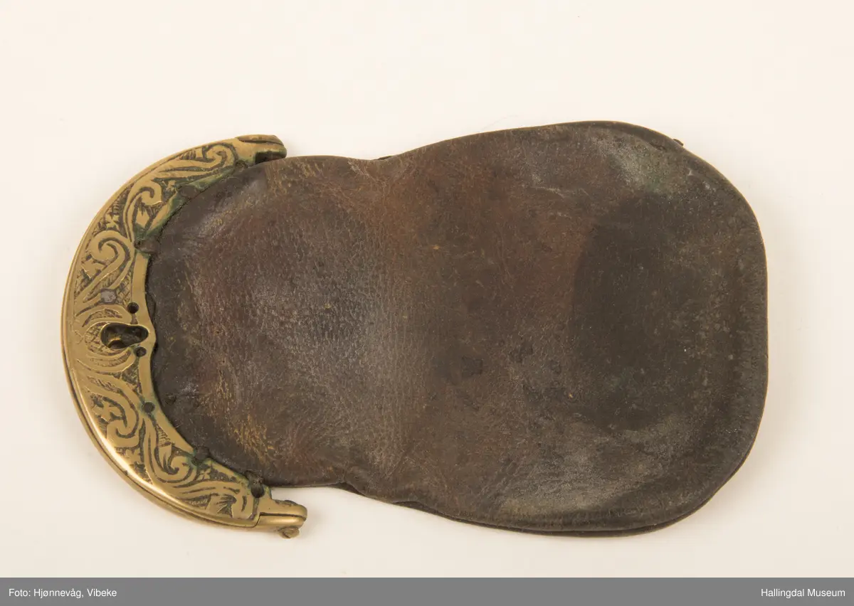 Lær, brunt. Beslag og lås av messing. Støpt. Sydd fast til pungen med lærbånd. Dekor. Akantusranke, gravert. L: 12,7 cm, B: 8 cm.
