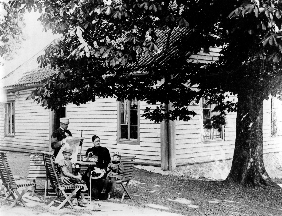 Familjen Appelbom sitter i trädgårdsmöbler framför huset. Viktor Appelbom var Ösmos komminister och de bodde i Nyble prästgård, som idag är Ösmos hembygdsgård. Viktor och hans fru Hedvig gifte sig och flyttade in i prästgården 1884. 

Läsning, sommar, fritid.

Personer på bilden:
Stående: komminister Viktor Appelbom, sittande från vänster: dottern Ingrid, hustrun Hedvig och sonen Einar.