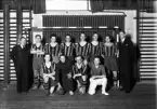 Foto av ett handbollslag med randiga tröjor, i en gymnastiksal. Vid sidorna om laget står ledarna.