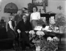 Foto av en kostymklädd man som sitter i ett vardagsrum, omgiven av sin familj och mängder av blommor.
50-årsdag (?).