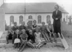 Foto av en skolklass med lärarinna, som samlats på trappan till en kyrkogård. I bakgrunden syns en kyrka.