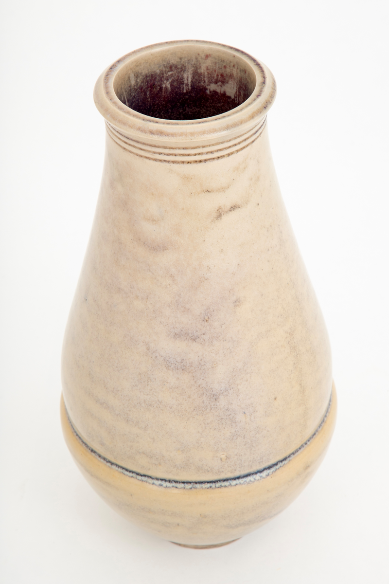 Vase i glasert steingods. Balusterformet korpus som hviler på en liten fotring. Overgangen mellom korpus og munningen er markert med tre horisontale linjer. Svakt utoverbrettet munningsrand. Vasens ytterside er dekket med en beigefarget glasur, mens innersiden har en rød-lilla fargetone.