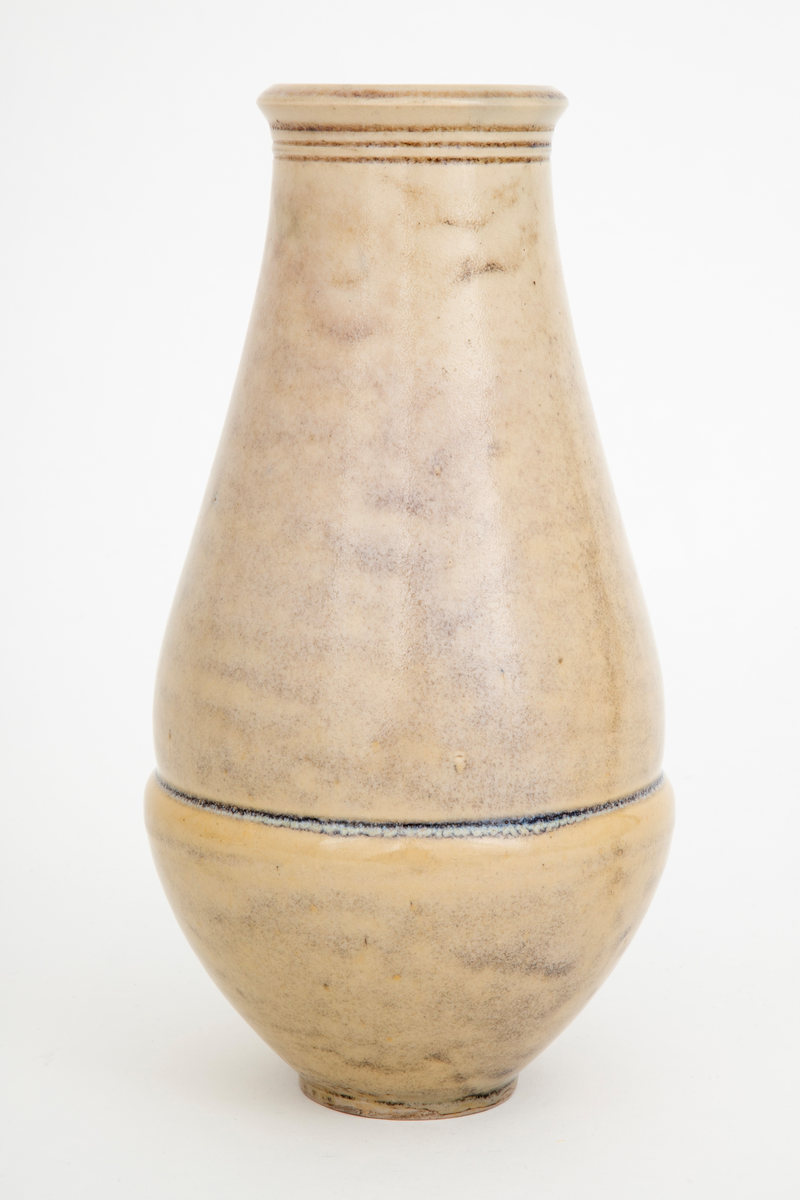 Vase i glasert steingods. Balusterformet korpus som hviler på en liten fotring. Overgangen mellom korpus og munningen er markert med tre horisontale linjer. Svakt utoverbrettet munningsrand. Vasens ytterside er dekket med en beigefarget glasur, mens innersiden har en rød-lilla fargetone.