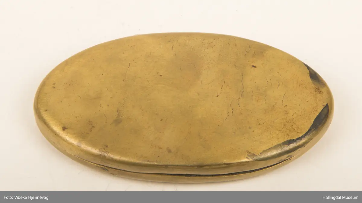 Messing, støpt. Oval, flat bunn, svakt hvelvet. Hengslet lokk. Ingen dekor. L: 9,3 cm, B: 5,3 cm.