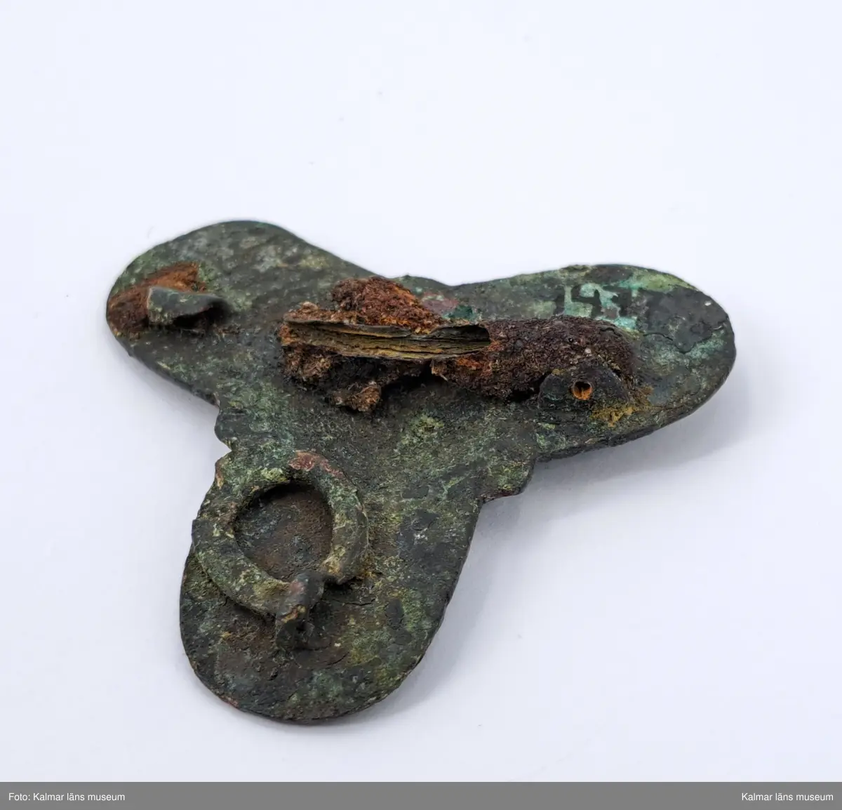 KLM 1477. Spänne, treflikigt spänne. Gjutet av brons. Snirklig ornamentik. På baksidan en ring för en kedja. Även nålfäste samt del av järnnål kvar på baksidan.