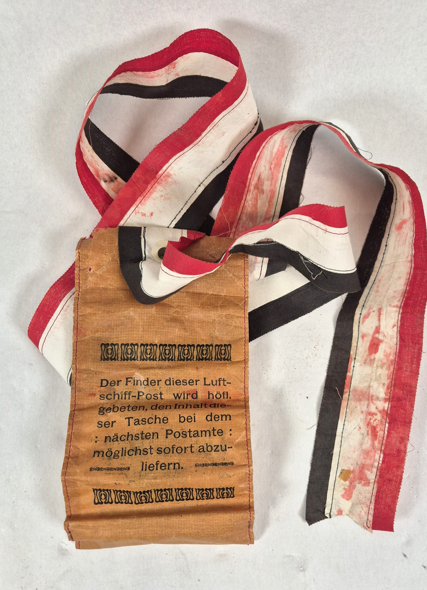 Pose med beskjed på tysk . Skrift og ornamentikk i Jugendstil (Art Nouveau)
Tekst: "Der Finder dieser Luftschiffpost wird höfl. gebeten den Innhalt dieser Tasche bei dem nächsten Postamte möglichst sofort abzuliefern".
Bånd er laget av tøy remser sydd sammen i fargene rød, hvit og svart.