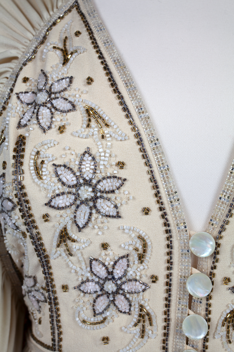 Fotsid selskapskjole i kremfarget silke med perlebrodert bærestykke med blomstermotiv. Dyp V-ringning og fire perlemorsknapper. Plisseringer under og rundt bærestykket og på skjørtet midt foran. Skjørtet er glatt bak og overlapper foran. Kjolen er uten ryggstykke og har i stedet for ermer to lange, draperte stoffstykker som kan lukkes i ryggen. Alle kantene har perlebroderte border.
