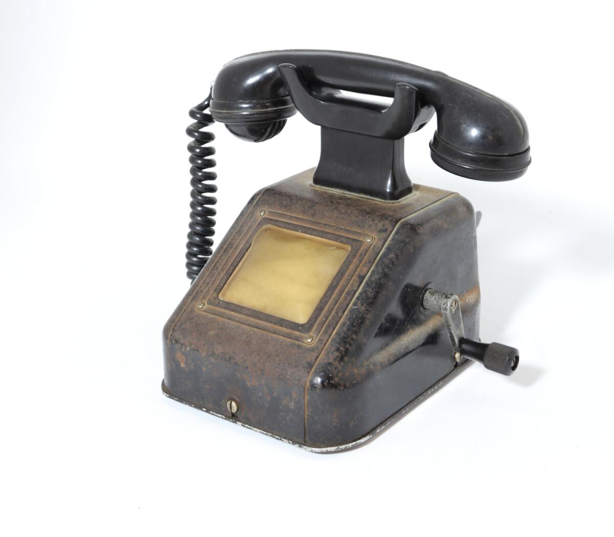 Telefonapparat for manuell sentral, brukt på setesdalsbanen. Apparatet har rektangulær bunn, rette sider, og skrå forkant med flat topp.  Det er en sveiv på siden, og ramme i front med plastdeksel.