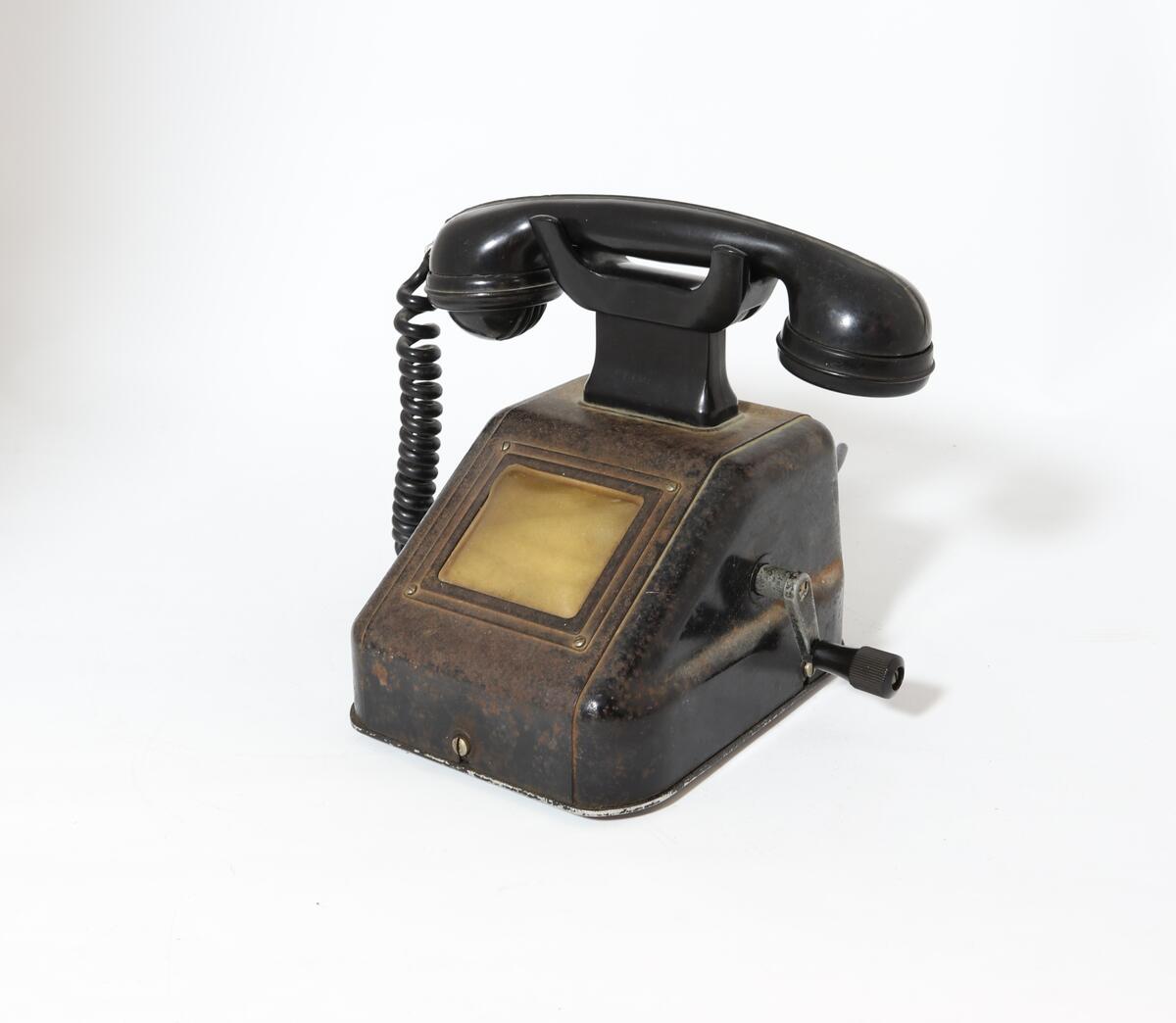 Telefonapparat for manuell sentral, brukt på setesdalsbanen. Apparatet har rektangulær bunn, rette sider, og skrå forkant med flat topp.  Det er en sveiv på siden, og ramme i front med plastdeksel.