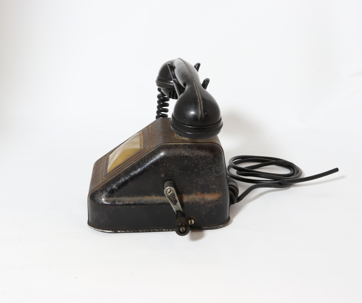 Telefonapparat for manuell sentral, brukt på setesdalsbanen. Apparatet har rektangulær bunn, rette sider, og skrå forkant med flat topp.  Det er en sveiv på siden, og ramme i front med plastdeksel.