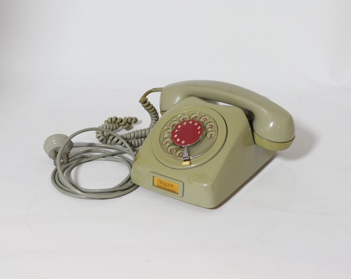 Gulnet grå telefon. Telefonen har tilnærmet rektangulær grunnflate og svakt buede sider med spor for telefonrøret. På fronten er det en talldreieskive.
