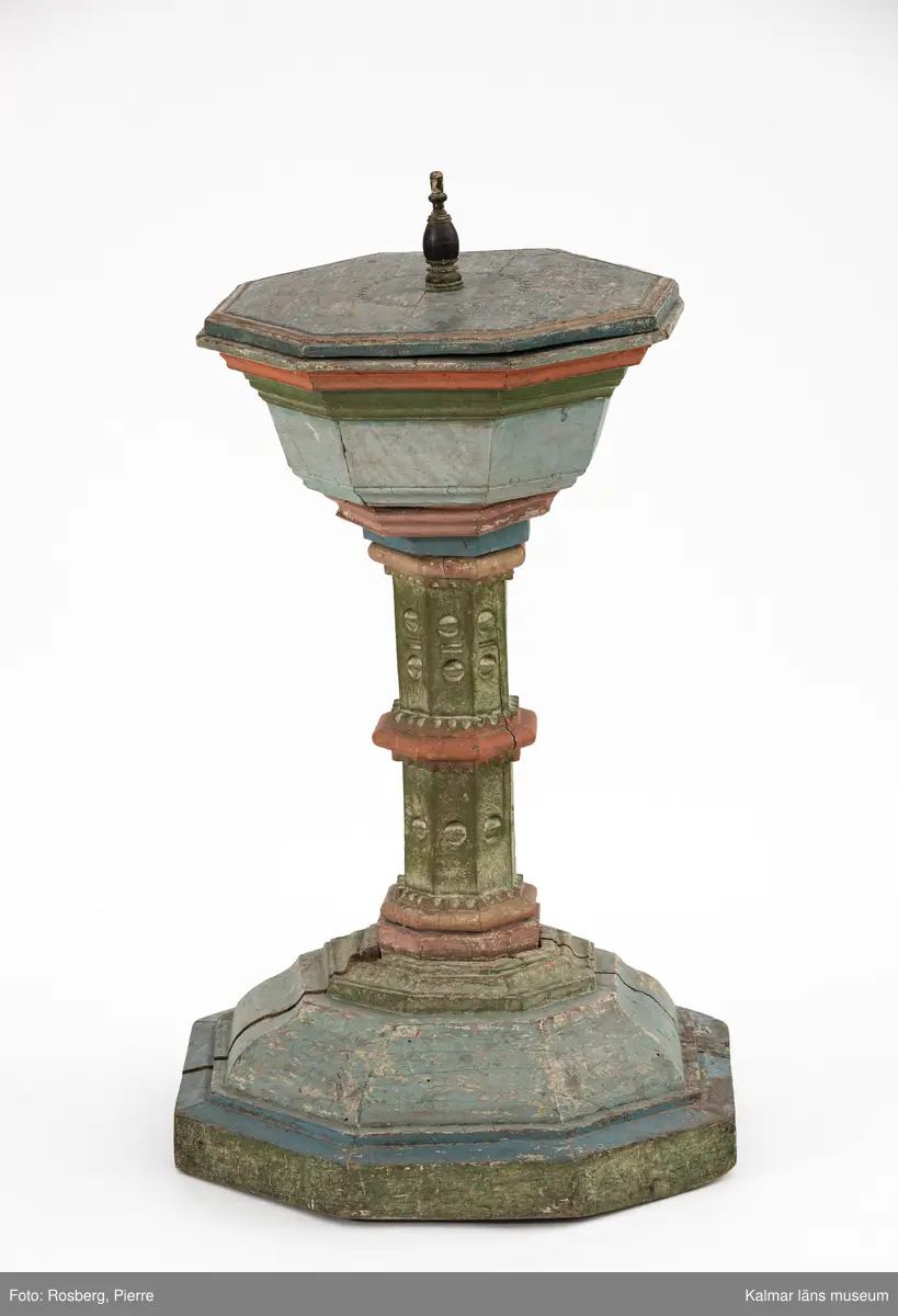 KLM 8072. Dopfunt. Av ek. Bemålad i blå, gröna och röda färger, locket är blåmålat med en svart knopp. Undertill syns vit färg med röd marmorering. Åttakantig form, profilerad och snidad pelare med nod. Vid åttakantig fot. Locket är även det åttakantigt, runt den svarvade knoppen finns skuren dekor med nagelsnitt. Plats för dopskål i funten.
