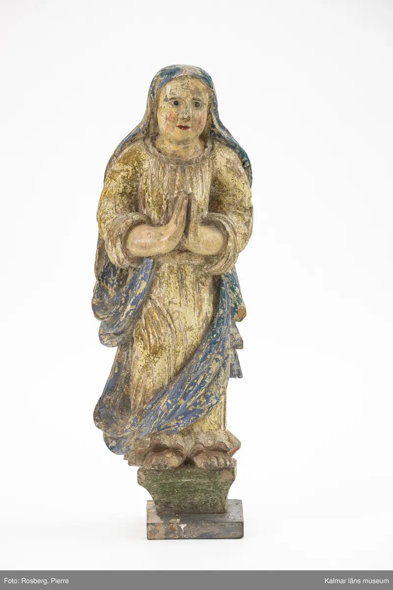 KLM 10488. Skulptur, kyrklig skulptur. Av trä. Föreställande en kvinnlig figur, eventuellt jungfru Maria. Kvinnan står barfota på en upphöjd bas. Hon har förgylld klänning med lång blå sjal som täcker hennes hår och draperas ned till fötterna.