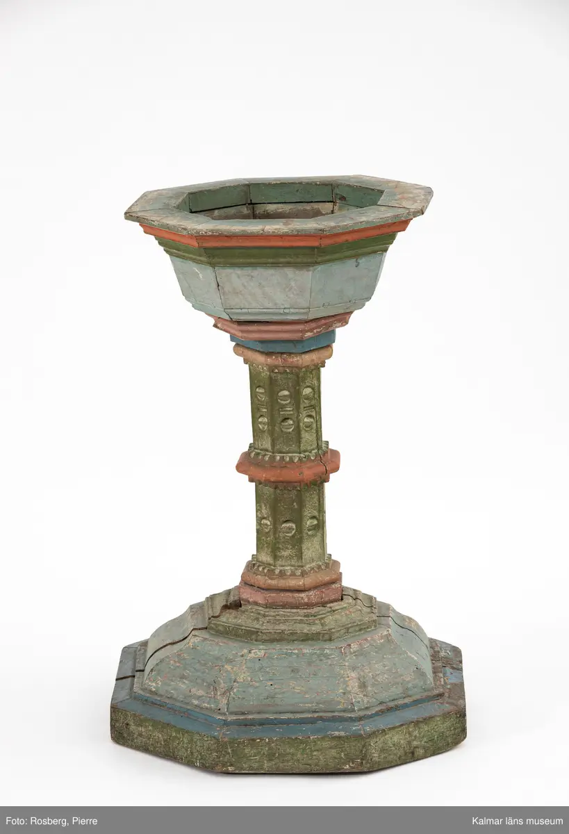 KLM 8072. Dopfunt. Av ek. Bemålad i blå, gröna och röda färger, locket är blåmålat med en svart knopp. Undertill syns vit färg med röd marmorering. Åttakantig form, profilerad och snidad pelare med nod. Vid åttakantig fot. Locket är även det åttakantigt, runt den svarvade knoppen finns skuren dekor med nagelsnitt. Plats för dopskål i funten.