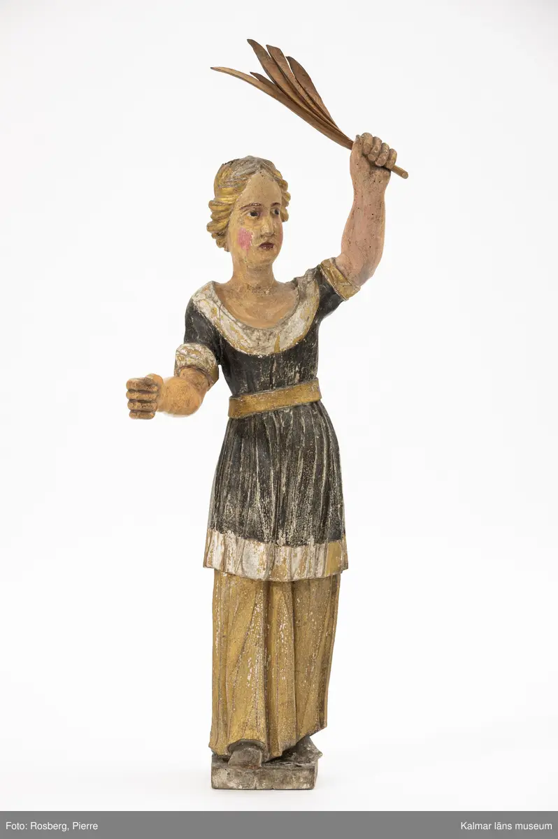 KLM 34771. Skulptur, kyrklig skulptur. Av trä. Föreställande en kvinnlig figur med en draperad klädsel målad i svart samt med förgyllning. Håret är uppsatt och förgyllt. Resturerad. I ena handen en palmkvist, omålad. Har ursprungligen hållit en fana i den lediga handen.
