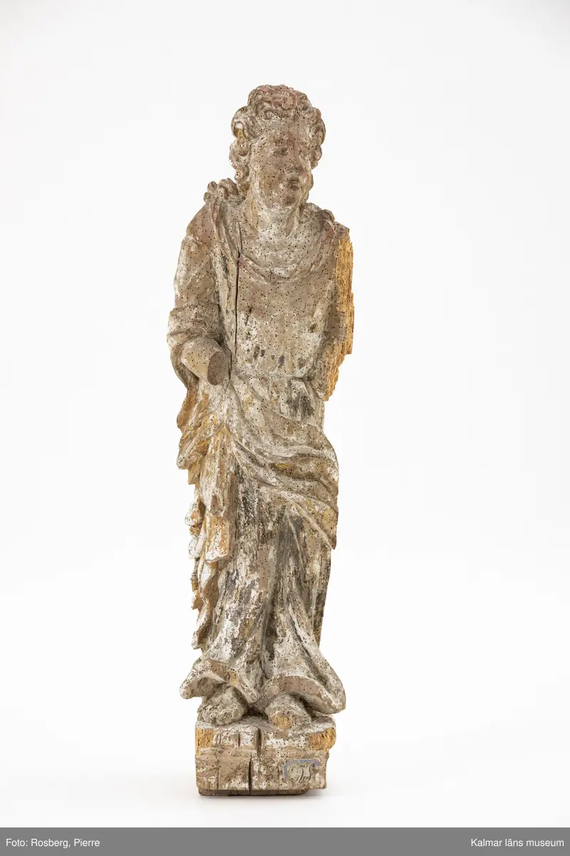 KLM 46859. Skulptur, kyrklig skulptur. Av trä. Skulptur föreställande en kvinnofigur med draperad fotsid klädsel och långt lockigt hår. Rester av grundering, färgskikt och förgyllning. Färgskikt i svart, grått, brunt, rött och grönt. Står barfota på en sockel. Skulpturen bär tydliga spår av tidigare skadedjursangrepp.