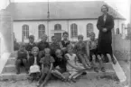 Foto av en skolklass med lärarinna, som samlats på trappan till en kyrkogård. I bakgrundens syns en kyrka.