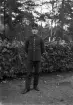 Foto av en man i mörk militäruniform, stående vid en häck.