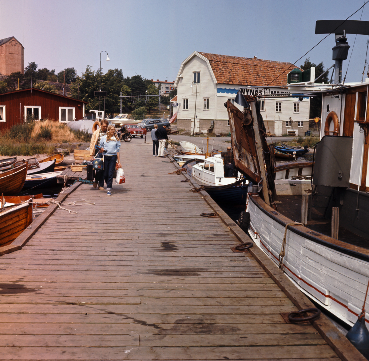 Hamnen, järnvägsbryggan med kyrkan och kvarnen i bakgrunden. Några personer med kassar i händerna är på väg ut på bryggan där småbåtar och fiskebåtar ligger förtöjda. 

Sjöfart, fritid, sommar.