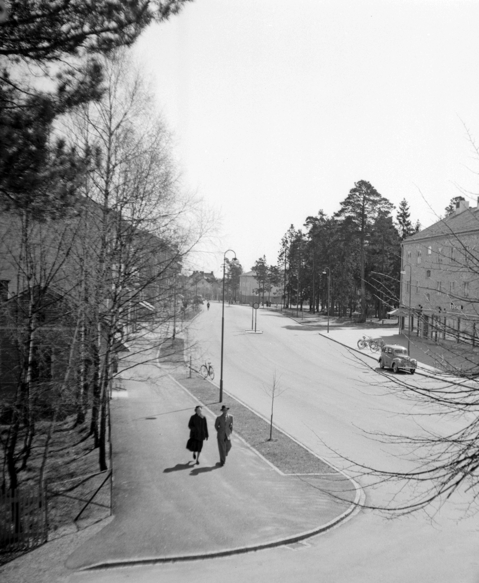 Vy över Nynäsvägen söderut från nr 38. Två personer går på trottoaren. På andra sidan gatan ser man huset som inrymde Konsum. En bil står parkerad vid trottaren. 

Vänstertrafik, handel.