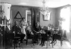 Foto av en familj i en salong. De blir just serverade kaffe av husan/jungfrun.
Möjligen foto ur Betty Larsons arkiv. Växjö (företrädare till Anton Persson som fotograf).
