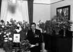 Foto av en man i kostym som sitter i ett vardagsrum,
omgiven av blommor.