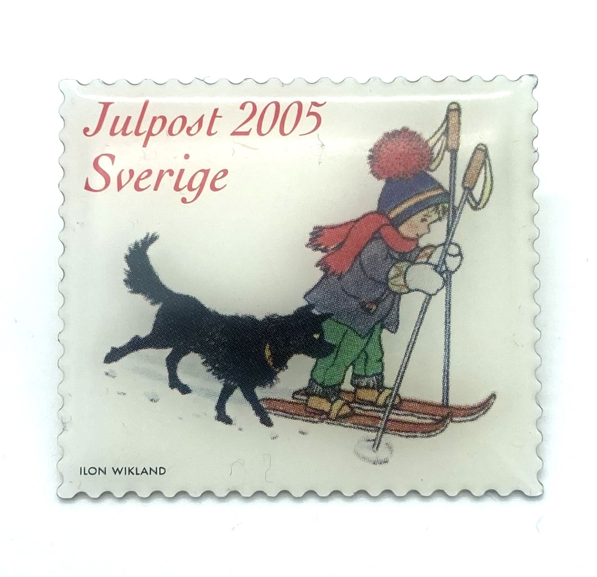 Pin i frimärksform med röd kursiverad text: Julpost 2005 Sverige. 
Tilll höger ett barn med mössa på med röd tofs blå hätta. Röd halsduk blå jacka gröna byxor, vita lovikavantar. Barnet åker skidor med två stavar och en svart hund följer med bredvid. Längst ner i vänstra hörnet signatur av illustratören ILON WIKLAND.