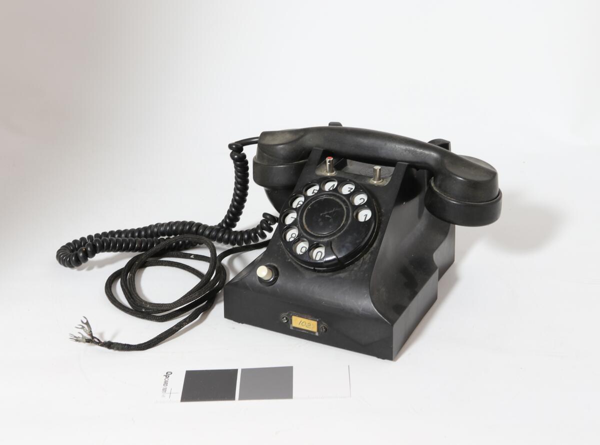 Vanlig bord-telefon med trykknapp under på siden av tallskiven. Telefonen har ekstra intern linje. Er et såkalt sekretærapparat. Telefonen er utstyrt med tekstilsnor til mikrofon og støpsel. To trykknapper ved gaffelen for intern linje.
