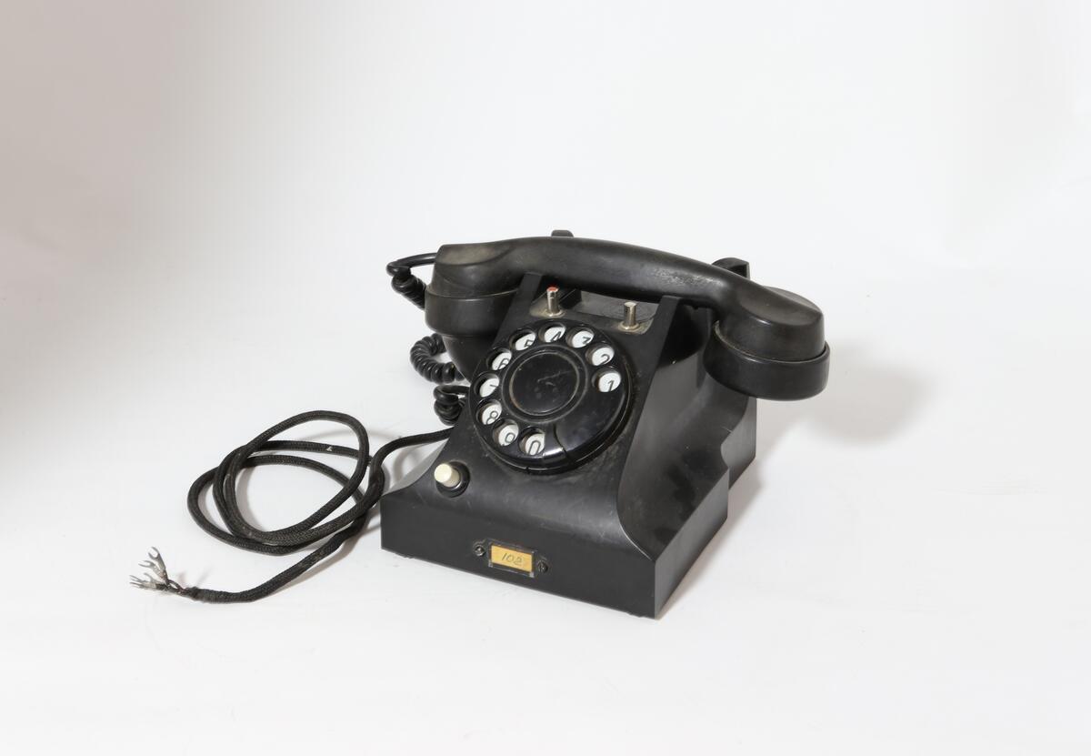 Vanlig bord-telefon med trykknapp under på siden av tallskiven. Telefonen har ekstra intern linje. Er et såkalt sekretærapparat. Telefonen er utstyrt med tekstilsnor til mikrofon og støpsel. To trykknapper ved gaffelen for intern linje.

