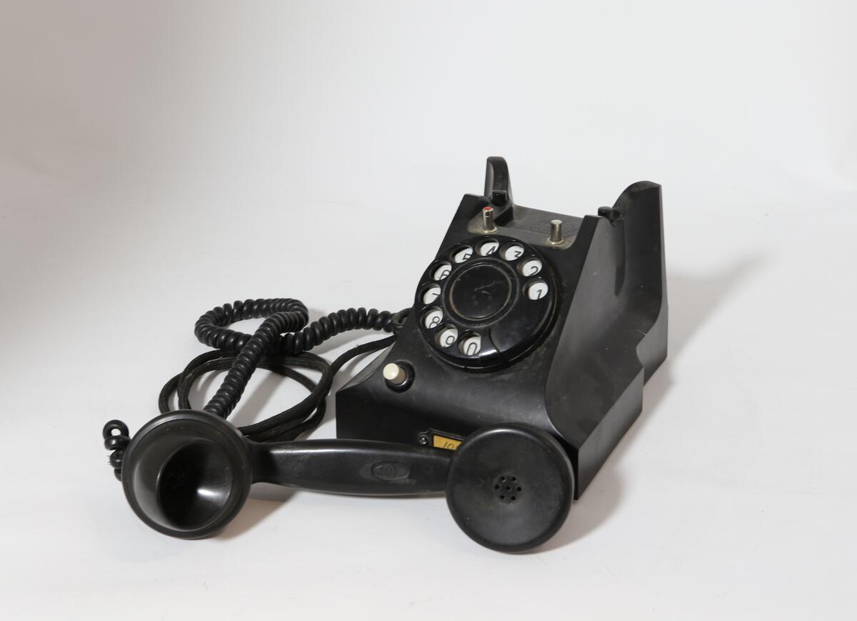 Vanlig bord-telefon med trykknapp under på siden av tallskiven. Telefonen har ekstra intern linje. Er et såkalt sekretærapparat. Telefonen er utstyrt med tekstilsnor til mikrofon og støpsel. To trykknapper ved gaffelen for intern linje.
